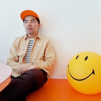 Photo of Jimmy Lee, sitting next to a yellow smiley face lamp in a cafe. He is wearing an orange cap, which is colour co-ordinated with the bench he is seated on, and looking away from the camera outside the window